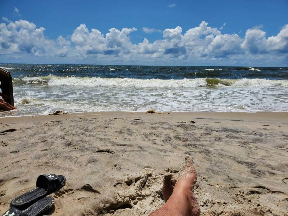 This image features a wide, sandy beach with gentle waves rolling in under a partly cloudy sky. The shoreline is open and inviting, providing ample space for classic beach activities like sandcastle building, beachcombing, and sunbathing.