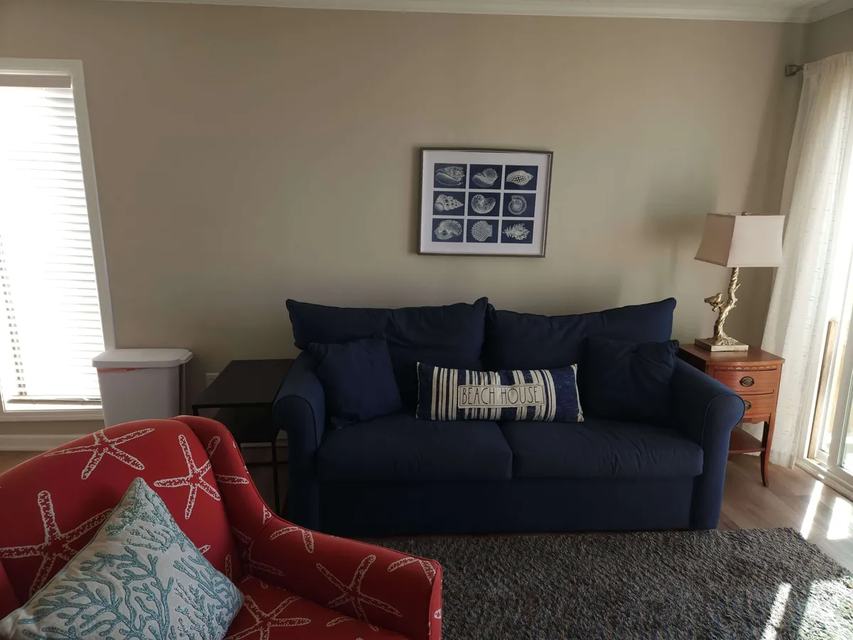 A cozy living area with a plush blue sofa, vibrant red accent chair, framed artwork, and warm lighting from a table lamp. The room feels inviting and tailored for relaxation or social connection.