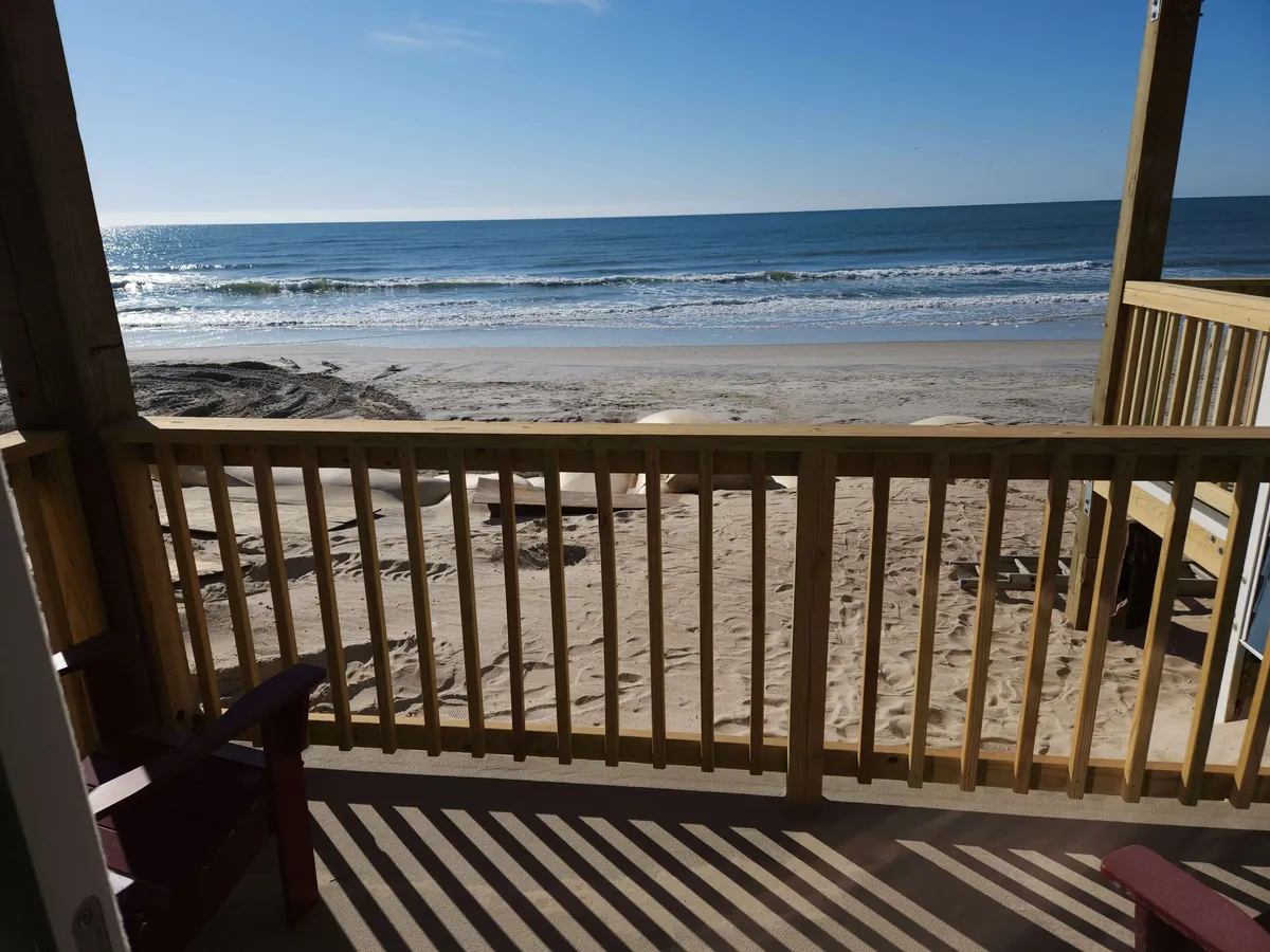 The hero image showcases a peaceful, oceanfront scene from the condo's private balcony. The view includes a wooden railing, comfortable deck chairs, and an uninterrupted vista of the beach and sparkling Atlantic Ocean beyond. The sand invites guests to build sandcastles or enjoy leisurely strolls, while the tranquil waves and possible dolphin sightings create a serene atmosphere.