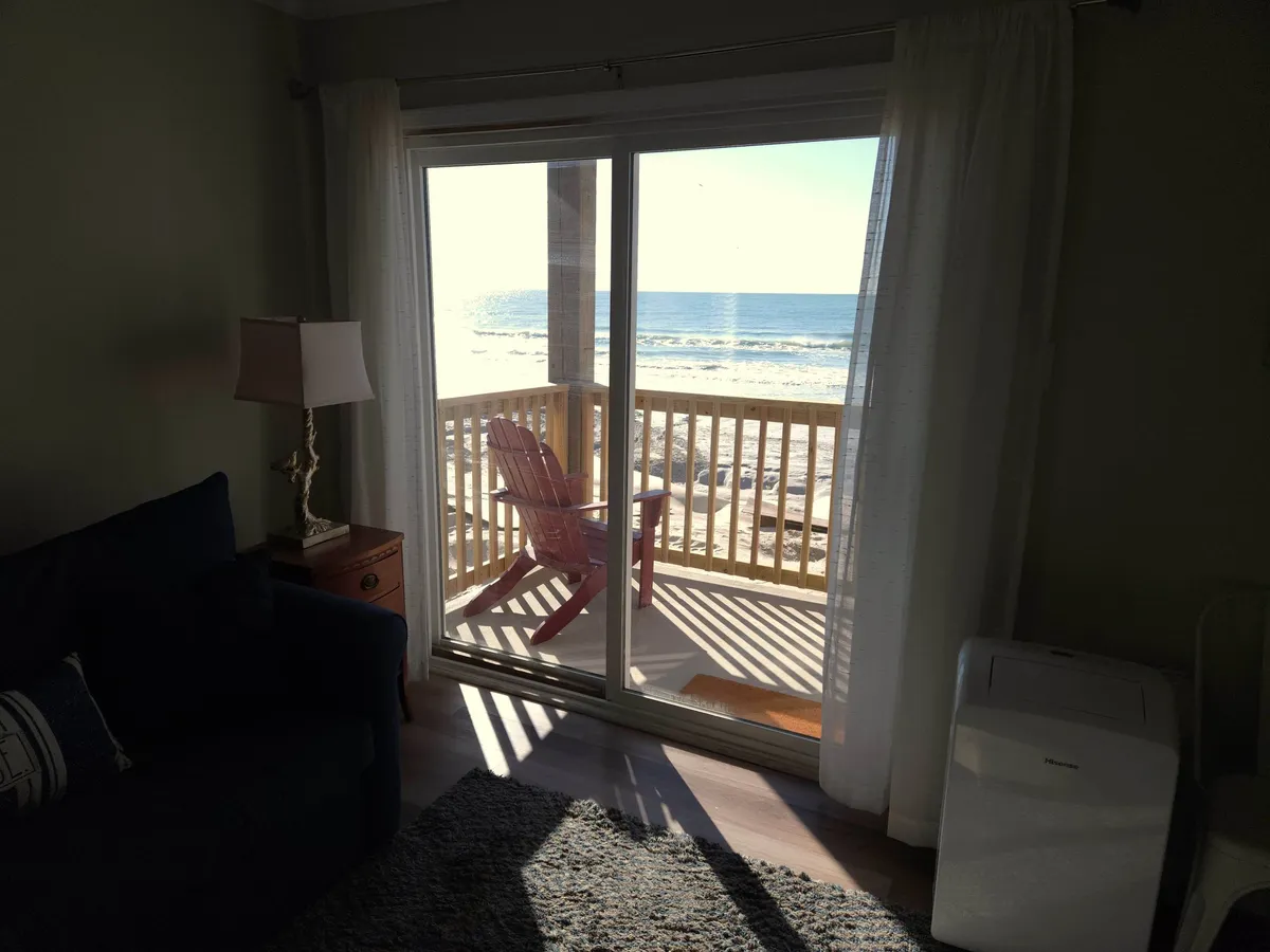 This image features a tranquil interior scene with a sliding glass door that opens directly to an ocean view. A red Adirondack chair sits beside the door, with a cozy sofa and lamp completing the welcoming entry sequence. Natural light pours in, highlighting the seamless flow from the entryway into the living space and drawing attention to the view beyond.