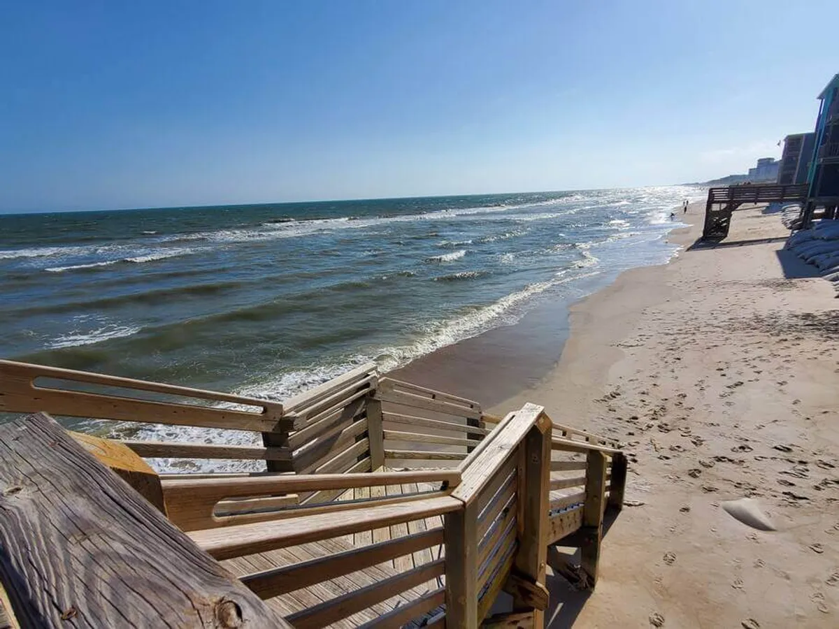 A wide, relaxed beach view featuring the shoreline, incoming ocean waves, and a boardwalk leading directly to the sand. The scene captures both the beauty of the natural surroundings and the inviting access to the beach, with a tranquil atmosphere perfect for leisurely moments.