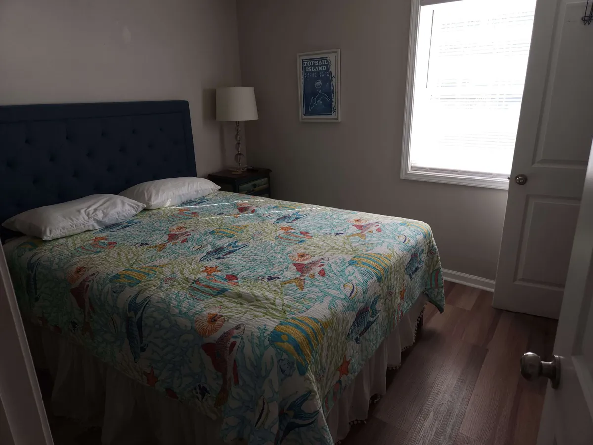 A tranquil bedroom featuring a bed with a colorful quilt, soft lamp lighting, and framed artwork on the walls. Natural light seeps in, adding to the calm and restful atmosphere.
