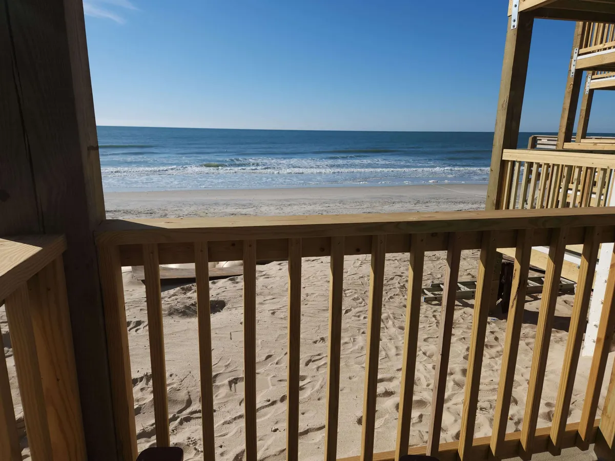 A stunning view from a private balcony overlooking the ocean, framed by a wooden railing. The sandy beach stretches out below, with gentle waves rolling in and the open water glimmering under the soft light. The vantage point perfectly captures the feeling of being perched right above the shore, offering both intimacy and awe-inspiring scenery.