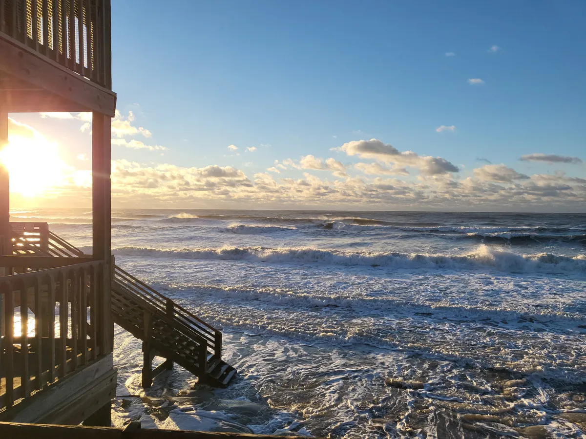 A serene sunset scene, with the sun setting on the horizon over the ocean, waves gently crashing on the shore, and a wooden staircase leading down to the beach. The soft evening light and calm mood evoke reflection and romance.