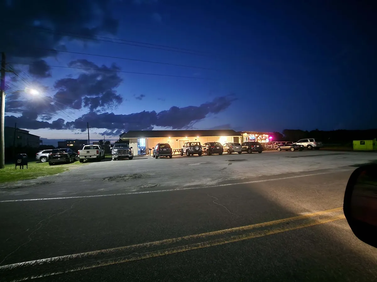 A lively evening exterior shot of a building with illuminated signage, parked cars, and glowing streetlights. The scene exudes a sense of community and activity, with people gathering for dining, socializing, and live entertainment.